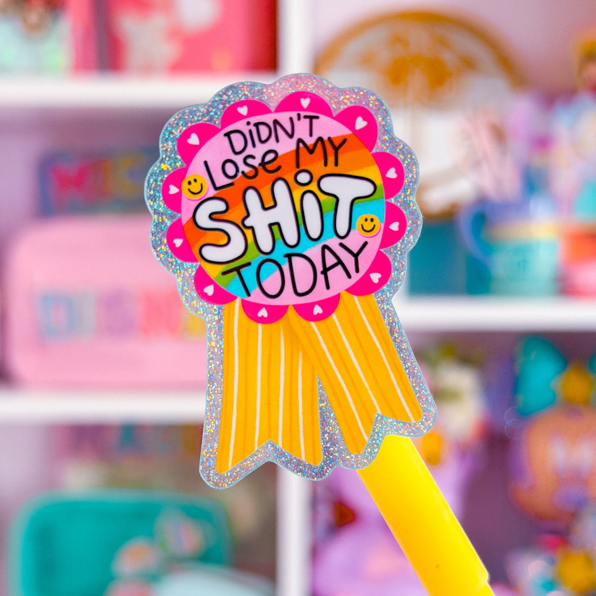 A close-up of a yellow pen with a colorful, glittery ribbon-shaped sticker that reads 'DIDN'T WANT TO LOSE MY SHIT TODAY' on a blurred pink and purple background.