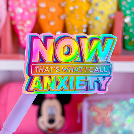 A colorful novelty sticker or sign reading 'NOW THAT'S WHAT I CALL ANXIETY' held in front of shelves of pink and multicolor glittery accessories.