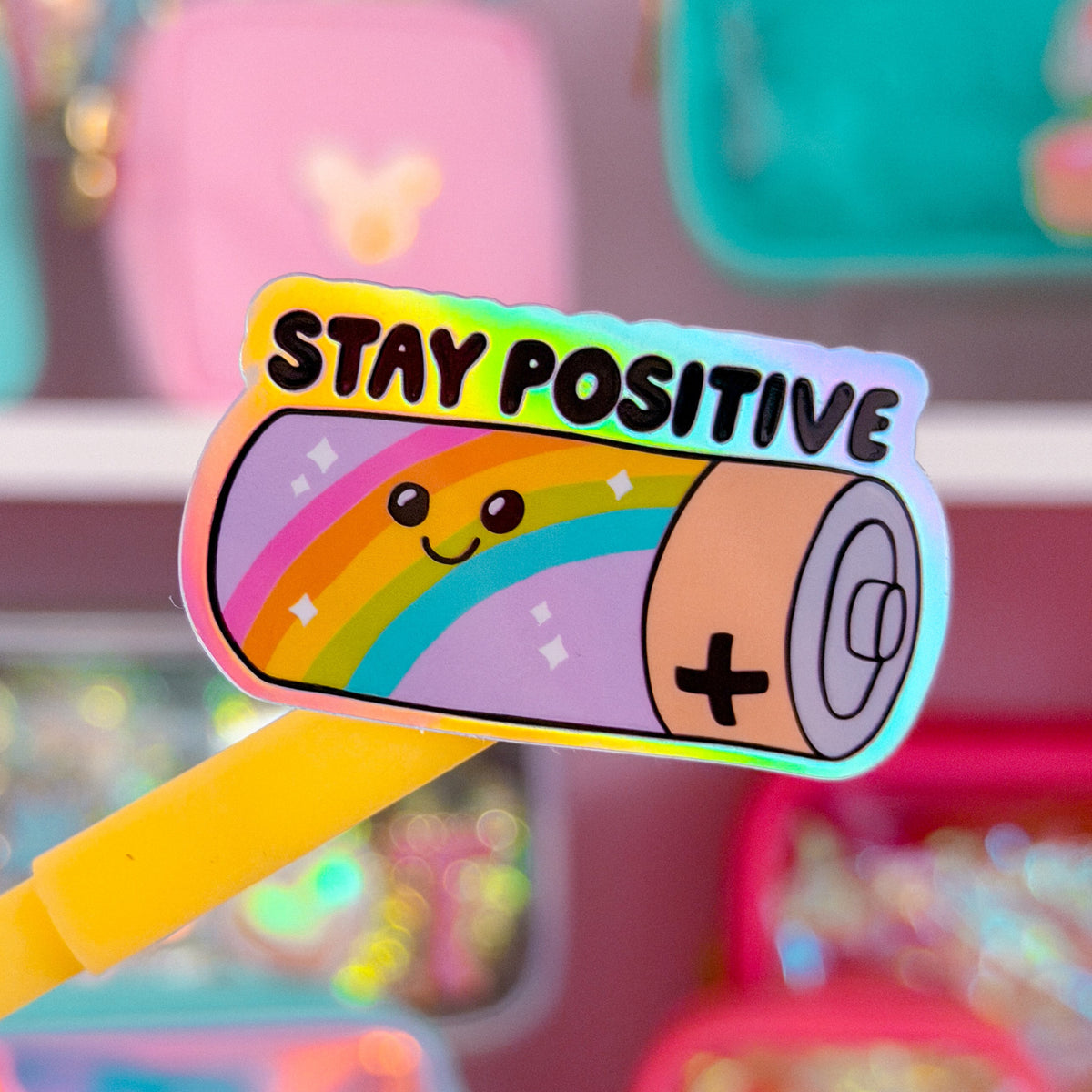 A close-up of a colorful sticker or patch featuring a sleeping cartoon character with a rainbow and the words “STAY POSITIVE,” held on a yellow stick against a blurred pastel shop background.