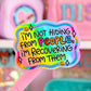 A close-up of a pink-handled sticker or card featuring a colorful wavy sticker that reads, 'I'm Not Hiding From People, I'm Recovering From Them' in playful multicolor lettering, with a blurred pink stationery or accessory display in the background.