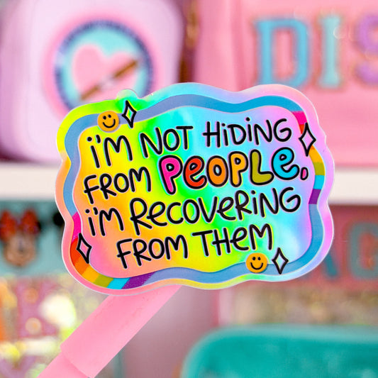 A close-up of a pink-handled sticker or card featuring a colorful wavy sticker that reads, 'I'm Not Hiding From People, I'm Recovering From Them' in playful multicolor lettering, with a blurred pink stationery or accessory display in the background.
