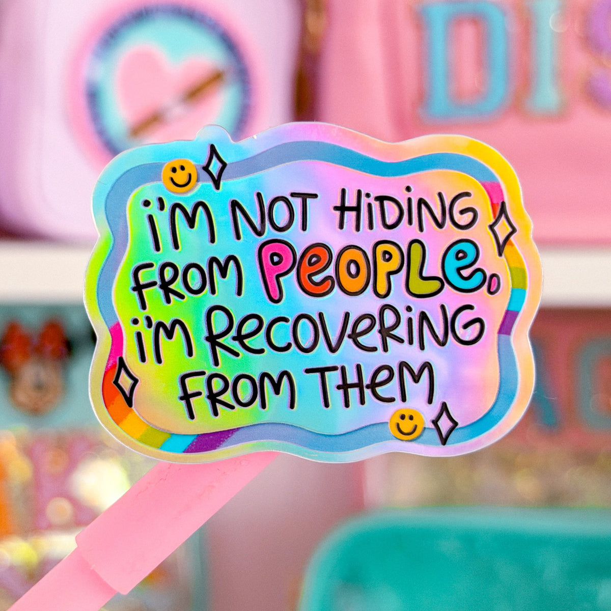 A pink-handled sticker or patch with a holographic cloud border and the text “I’m Not Hiding From People, I’m Recovering From Them,” shown in front of a blurred pink stationery or accessory display.
