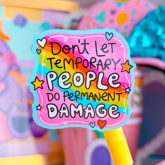 A colorful sticker on a yellow stick that reads 'Don't Let Temporary People Do Permanent Damage' in playful hand-drawn lettering, set against a blurred pastel toy background.