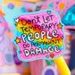 A close-up of a colorful sticker or label on a yellow stick, reading 'Don't Let Temporary People Do Permanent Damage' in playful hand-drawn lettering. The background is a soft-focus pastel scene with pink and purple objects.