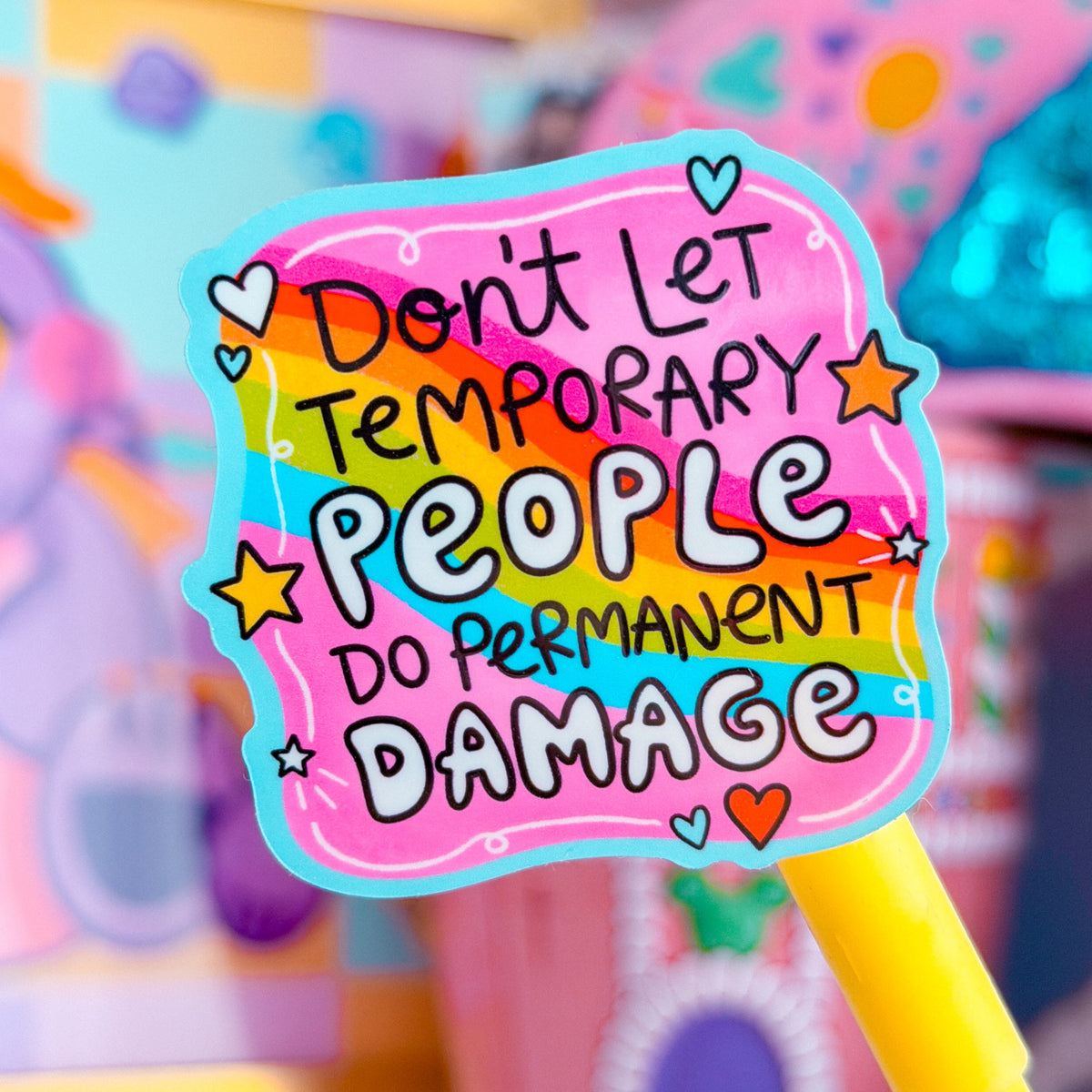 A close-up of a colorful sticker or label on a yellow stick, reading 'Don't Let Temporary People Do Permanent Damage' in playful hand-drawn lettering. The background is a soft-focus pastel scene with pink and purple objects.