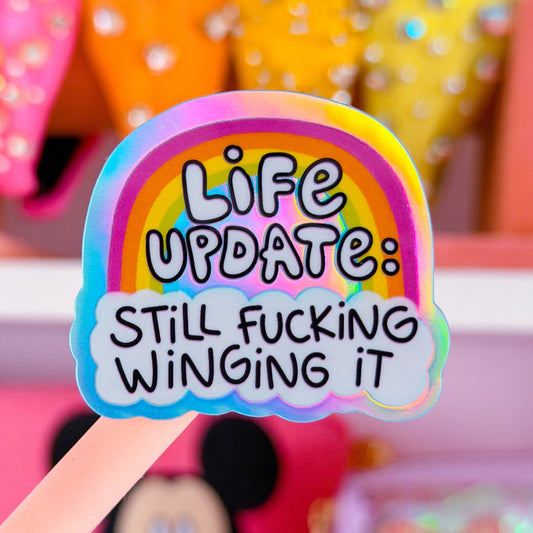 A close-up of a pastel rainbow sticker held on a pink stick, reading 'Life Update: Still Fucking Winging It,' with a blurred pink and multicolored background featuring rhinestone-covered items and a Mickey Mouse face.