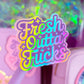 A pastel pink and purple novelty pen or stick with a large holographic sticker reading 'Fresh Outta Fucks' in rainbow gradient lettering, shown in a pink-themed setting with glittery packaging and a pink container in the background.