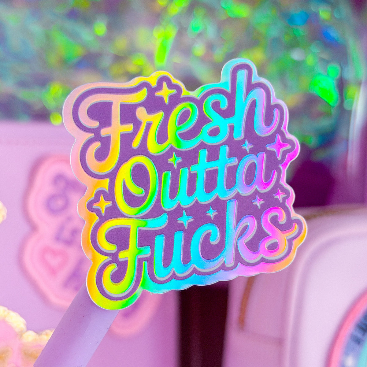 A pastel pink and purple novelty pen or stick with a large holographic sticker reading 'Fresh Outta Fucks' in rainbow gradient lettering, shown in a pink-themed setting with glittery packaging and a pink container in the background.