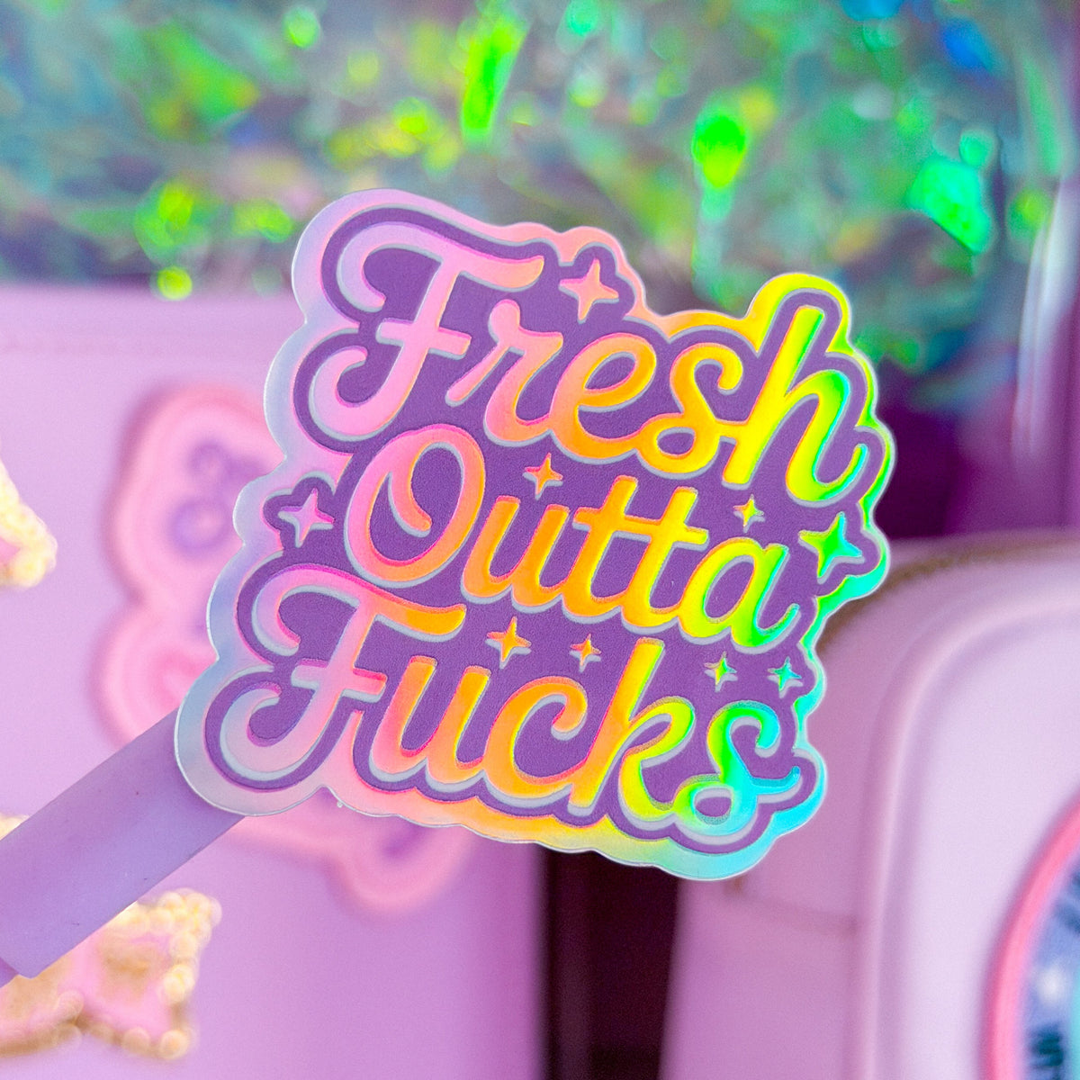 A close-up of a pastel pink pen with a holographic sticker that reads 'Fresh Outta' Fucks' in pink, purple, yellow, and blue gradient lettering. The background shows a matching lavender desk organizer with glittery compartments.
