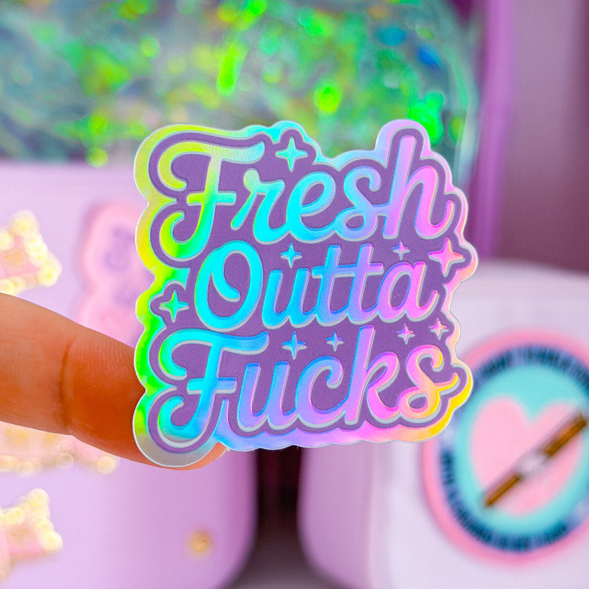 A close-up of a hand holding a colorful sticker that reads 'Fresh Outta Fucks' in a pastel gradient with holographic glitter accents, set against a blurred pastel desk background with stationery items.