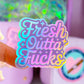 A close-up of a finger holding a holographic sticker that reads 'Fresh Outta Fucks' in pastel rainbow colors, with a blurred pink stationery or craft shelf in the background.