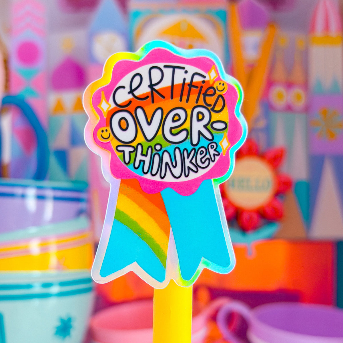A colorful 'Certified Over Thinker' ribbon-shaped sticker on a yellow stick, displayed in front of a blurred play kitchen scene with pastel toy cups and towers.