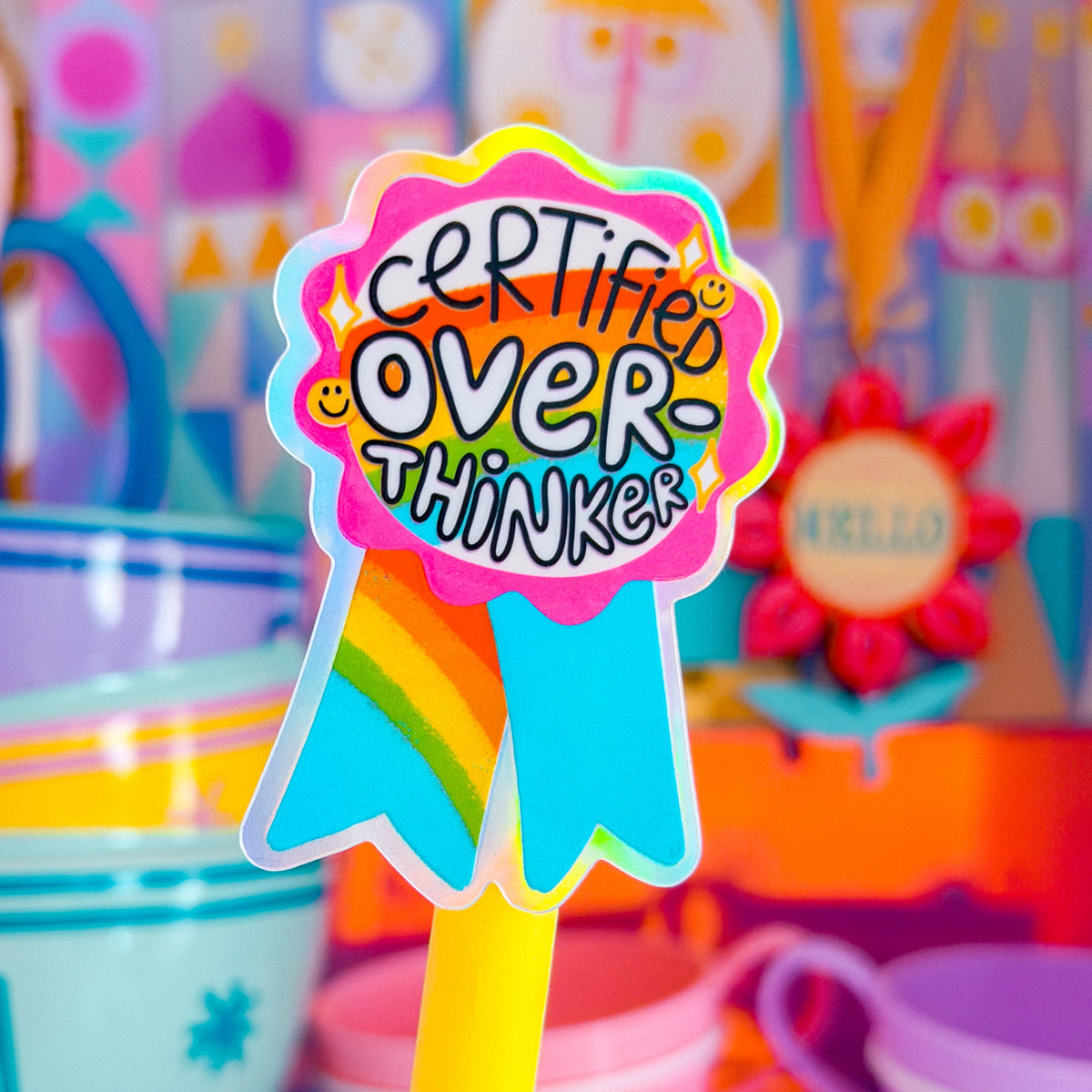 A colorful novelty pen topper or sticker reading 'CERTIFIED OVER THINKERS' in playful multicolor lettering, mounted on a yellow pen-like stick, with a blurred pastel toy-tea scene in the background.