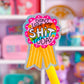 A close-up of a yellow pen with a glittery, rainbow-colored ribbon award badge that reads 'DIDN'T LOSE MY SHIT TODAY' against a blurred pink and purple toy or stationery shelf background.