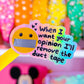 A novelty yellow-handled pen or pencil with a die-cut sticker that reads 'When I want your opinion I'll remove the duct tape' over a yellow smiley face with duct-taped lips. The item is shown in a colorful, blurred room setting with pink shelves and decorative objects in the background.