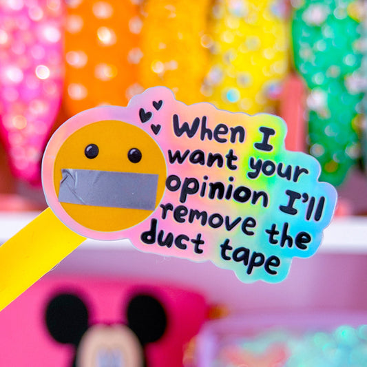 A novelty yellow-handled pen or pencil with a die-cut sticker that reads 'When I want your opinion I'll remove the duct tape' over a yellow smiley face with duct-taped lips. The item is shown in a colorful, blurred room setting with pink shelves and decorative objects in the background.