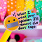 A yellow novelty pen or stick with a round emoji face taped mute and the text 'When I want your opinion I'll remove the duct tape' in the center, set against a blurred pink room background with colorful objects.