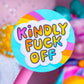 A close-up of a round pink lollipop-style sign or sticker on a pink stick, with the phrase “Kindly Fuck Off” in colorful bubble letters on a white cloud background. The scene is softly blurred with pastel pink and rainbow-colored objects in the background.