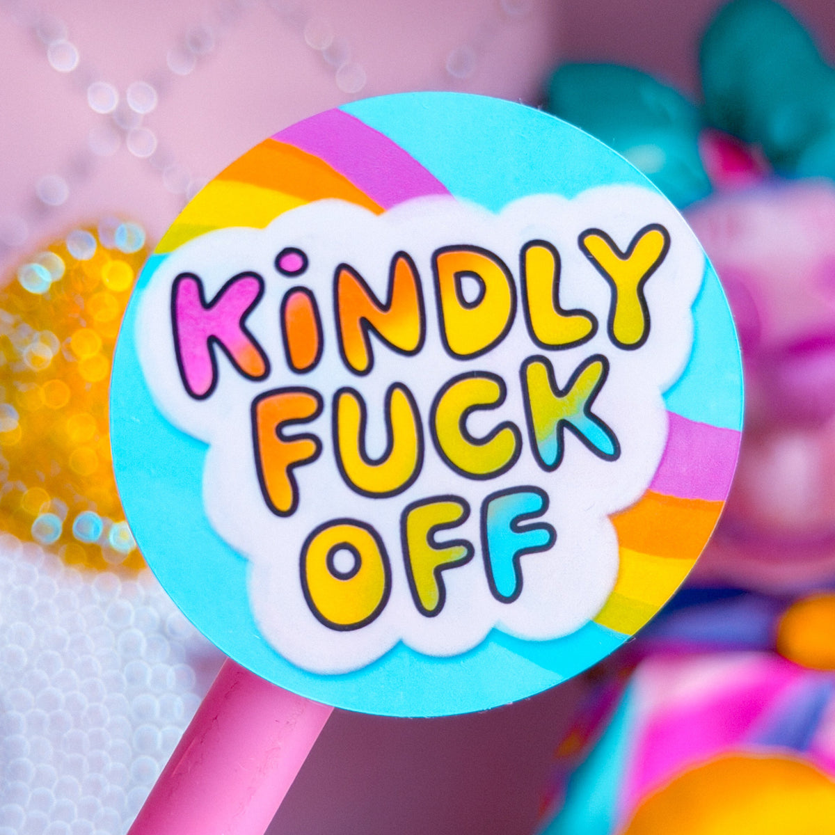 A close-up of a round pink lollipop-style sign or sticker on a pink stick, with the phrase “Kindly Fuck Off” in colorful bubble letters on a white cloud background. The scene is softly blurred with pastel pink and rainbow-colored objects in the background.