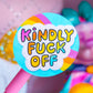 A close-up of a round sticker on a pink stick, reading 'Kindly Fuck Off' in colorful bubble letters. The sticker is in sharp focus, with a pink quilted bag and colorful plush toys blurred in the background.