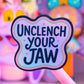 A close-up of a pink novelty pen or stick with a wavy sticker that reads “UNCLENCH YOUR JAW” in dark blue lettering. The background is a soft-focus pink and purple display with plush toys or decorative items.