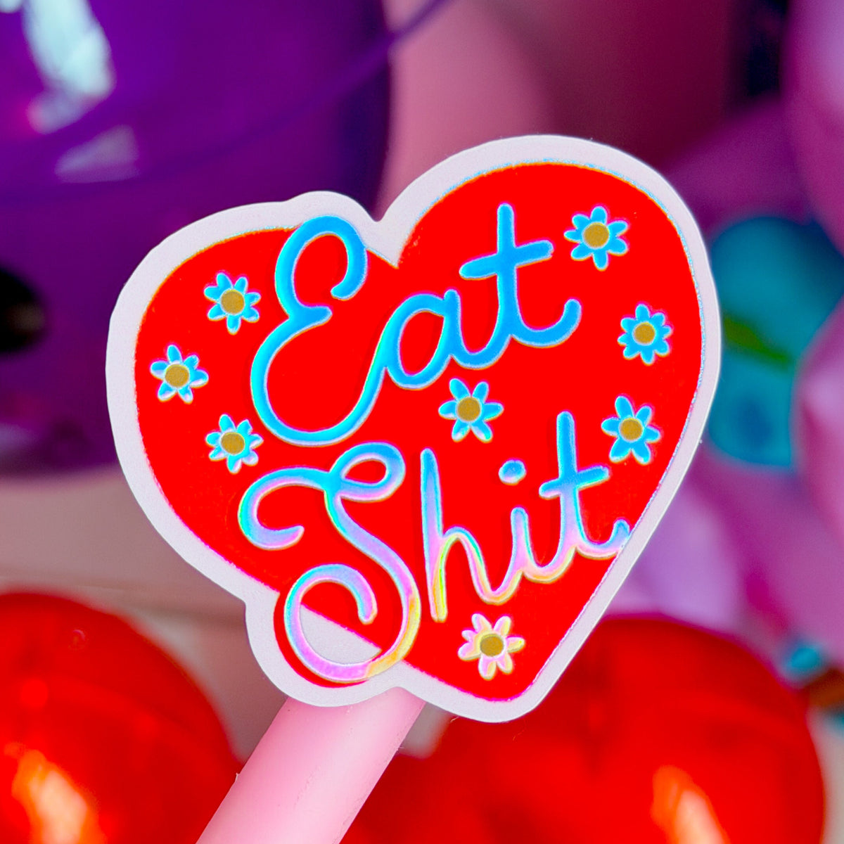 A close-up of a pink novelty lollipop or pen-style candy with a heart-shaped red top that reads 'Eat Shit' in blue script, surrounded by small white and blue flowers. The background is softly blurred with purple and pink objects.
