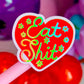 A heart-shaped novelty lollipop or sticker on a pink stick, featuring the phrase 'Eat Shit' in colorful neon-style lettering on a red background with small flower motifs.