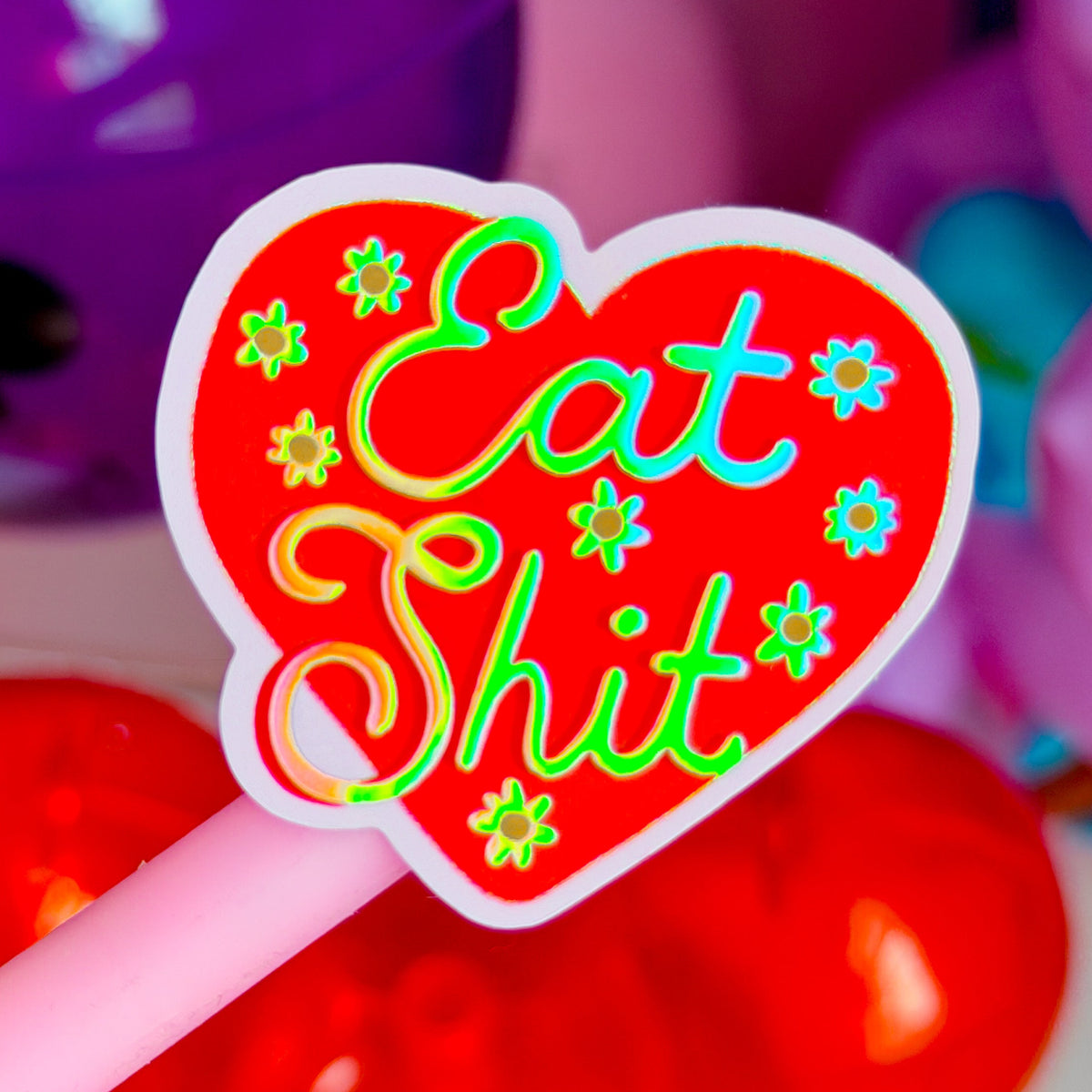 A heart-shaped novelty lollipop or sticker on a pink stick, featuring the phrase 'Eat Shit' in colorful neon-style lettering on a red background with small flower motifs.