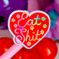 A pink-handled novelty lollipop or wand with a heart-shaped red sticker that reads 'Eat Shit' in colorful script, surrounded by small flower graphics, photographed against a blurred pink and purple background.