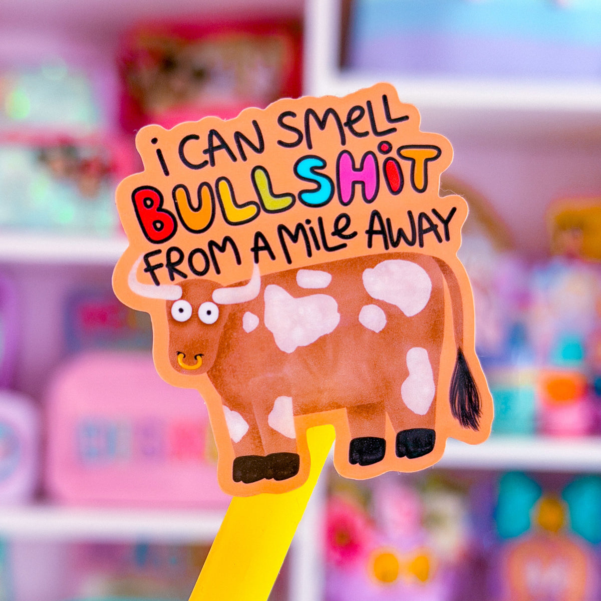 A novelty pen topper featuring a cartoon cow and the text 'i CAN SMELL BULLSHIT FROM A MILE AWAY' in colorful lettering, shown on a yellow pen against a blurred pink toy store background.