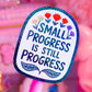 A close-up of a pink stick-shaped product with a glittery blue-edged label that reads “SMALL PROGRESS IS STILL PROGRESS” in dark blue lettering, decorated with small colorful flowers and leaves. The background is a soft pink, blurred scene with decorative objects.