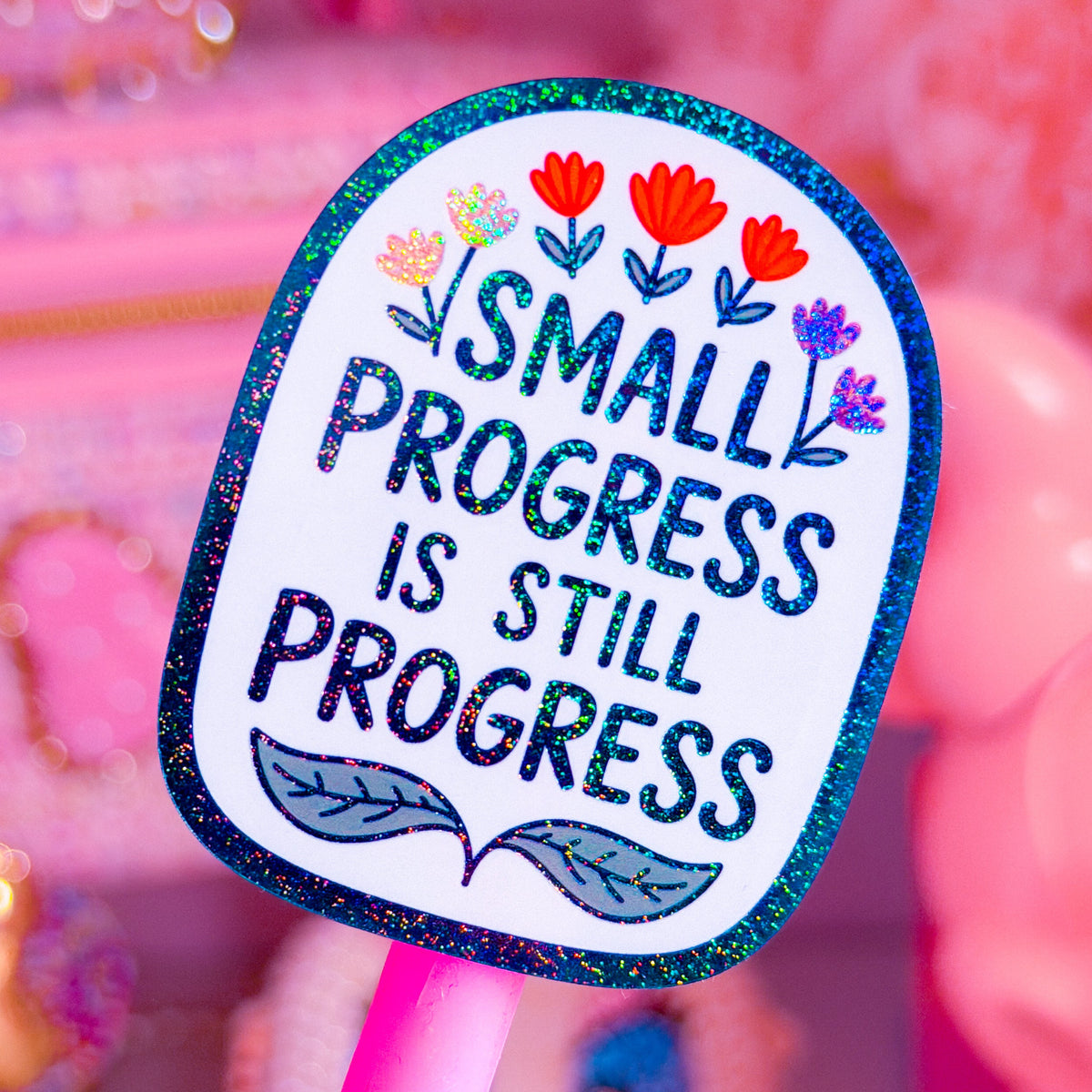 A close-up of a pink stick-shaped product with a glittery blue-edged label that reads “SMALL PROGRESS IS STILL PROGRESS” in dark blue lettering, decorated with small colorful flowers and leaves. The background is a soft pink, blurred scene with decorative objects.