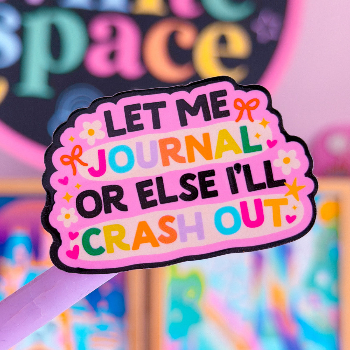 A close-up of a pink pen with a decorative sticker that reads 'LET ME JOURNAL OR ELSE I'LL CRASH OUT' in colorful lettering. The background is a soft-focus pink and multicolored setting with framed art and a sign reading 'white space'.