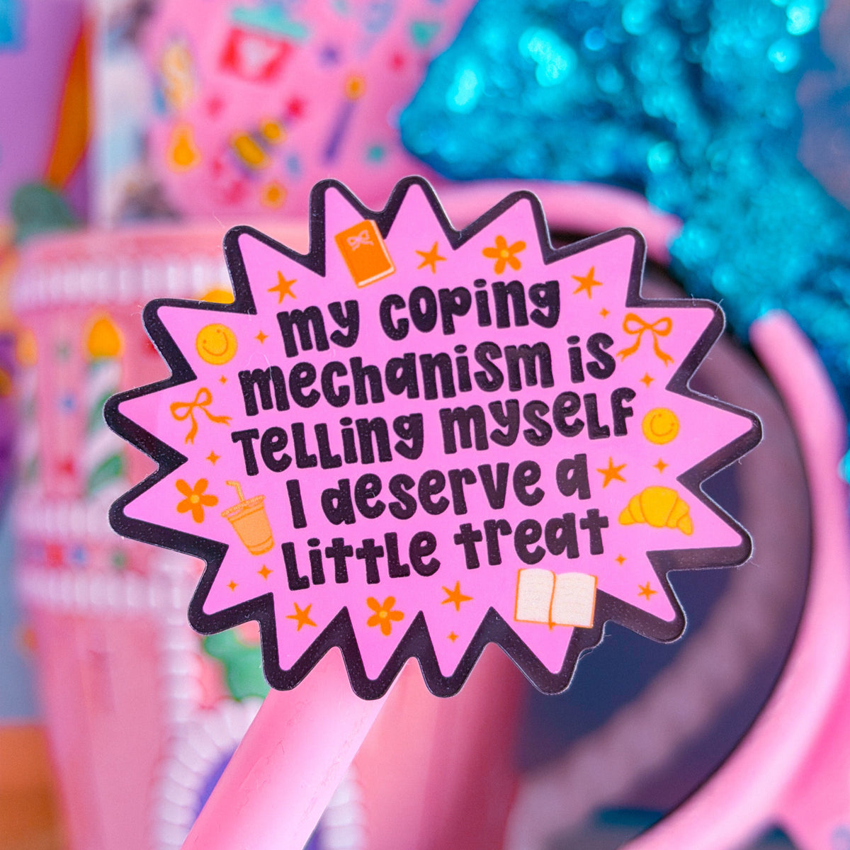 A pink novelty pen with a starburst-shaped pink end that reads, 'my coping mechanism is telling myself I deserve a little treat' in black text. The pen is shown in a colorful, pastel-themed setting with a pink headband, a glittery blue bow, and other pastel accessories in the background.