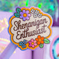 A close-up of a lavender pen with a decorative die-cut sticker that reads 'Shenanigan Enthusiast' in brown script, surrounded by colorful flowers. The pen is shown in a styled desk setup with a purple pencil case and other stationery items in the background.