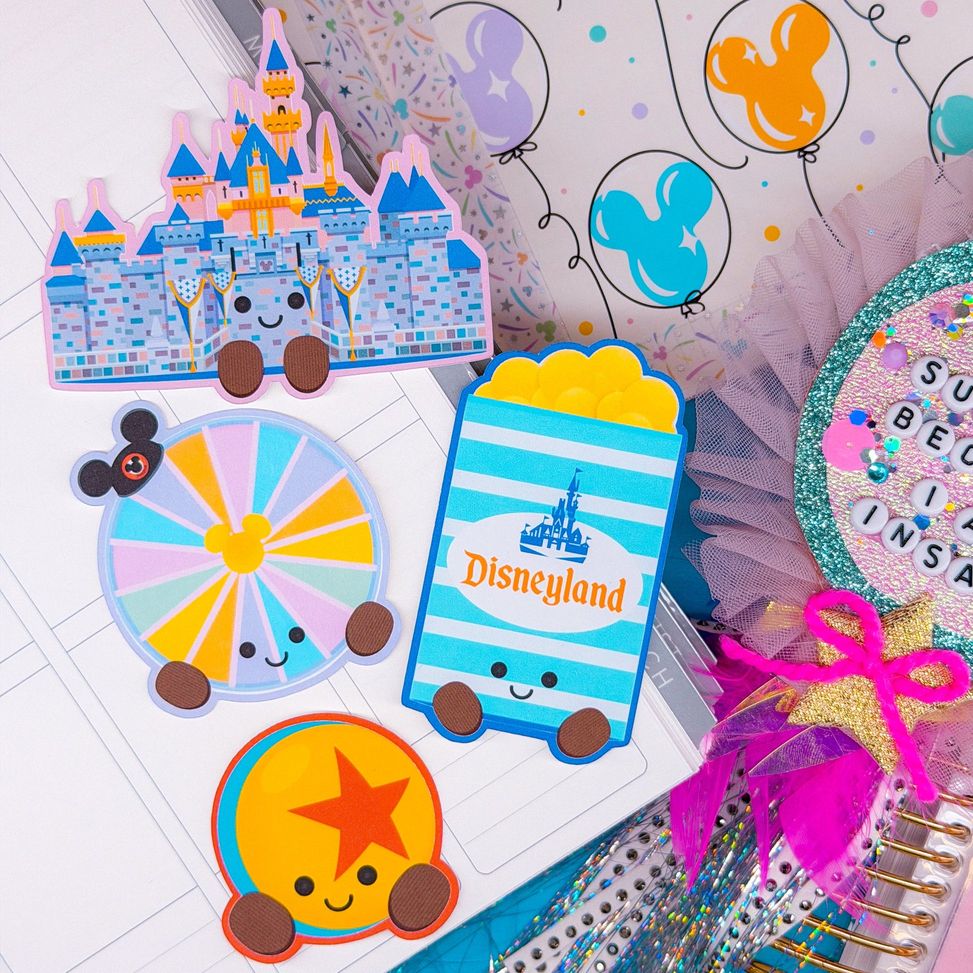 A styled flat-lay of Disney-themed stationery and sticker accessories, including a castle-shaped sticker, a popcorn box sticker with Disneyland branding, a Mickey Mouse wheel sticker, and a glittery circular embellishment with letter tiles, arranged on a white notebook and decorative paper.