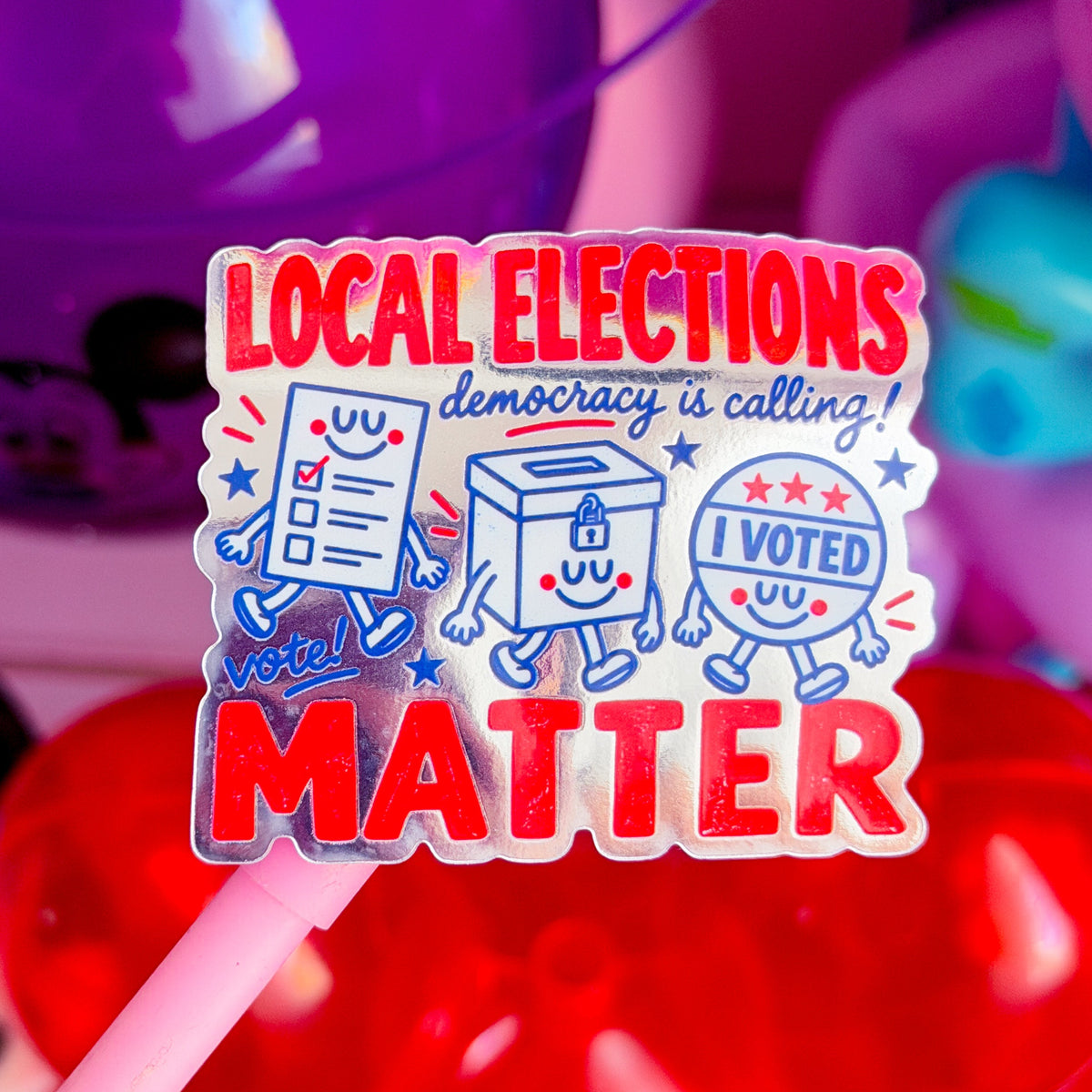 A die-cut sticker on a pink stick that reads 'LOCAL ELECTIONS democracy is calling! I VOTED MATTER' with cartoon ballot, box, and ballot-box characters. The sticker is photographed in a colorful, blurred setting with purple and red balloons in the background.