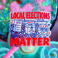 A close-up of a pink-handled sticker or card featuring the text 'LOCAL ELECTIONS democracy is calling! MATTER' with cartoon voting symbols in red, white, and blue, set against a colorful blurred background.