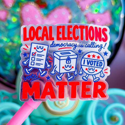 A close-up of a pink-handled sticker or card featuring the text 'LOCAL ELECTIONS democracy is calling! MATTER' with cartoon voting symbols in red, white, and blue, set against a colorful blurred background.