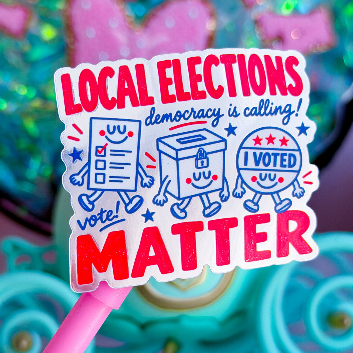 A close-up of a pink-handled sticker featuring cartoon ballot, envelope, and sign illustrations with the text 'LOCAL ELECTIONS democracy is calling! I VOTED MATTER' in red and blue on a white background.