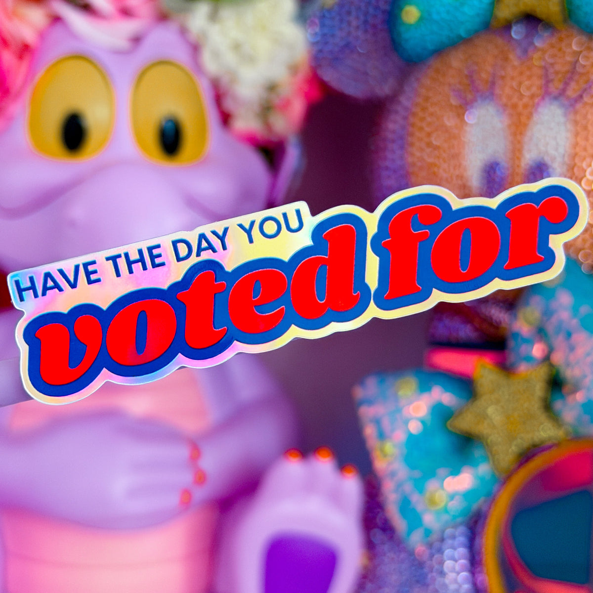A close-up of a colorful plush toy with a purple body, large yellow eyes, and a flower crown, alongside other bright stuffed animals and sequined accessories. A large text overlay reads 'HAVE THE DAY YOU voted for'.