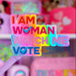 A close-up of a pink and multicolor sticker or decal that reads 'I AM WOMAN WATCH ME VOTE' with a heart and checkmark design, held in front of a blurred pink shelf background containing Disney-themed items.