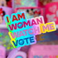 A close-up of a pastel purple pen with a colorful sticker that reads 'I AM WOMAN WATCH ME VOTE' in bold rainbow letters, set against a blurred pink background of shelves filled with stationery and Disney-themed items.
