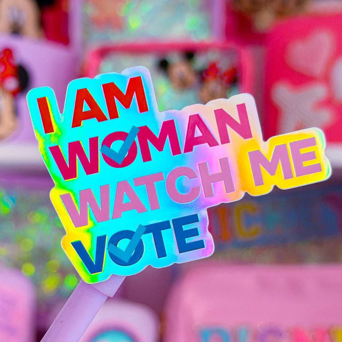 A close-up of a pastel purple pen with a colorful sticker that reads 'I AM WOMAN WATCH ME VOTE' in bold rainbow letters, set against a blurred pink background of shelves filled with stationery and Disney-themed items.