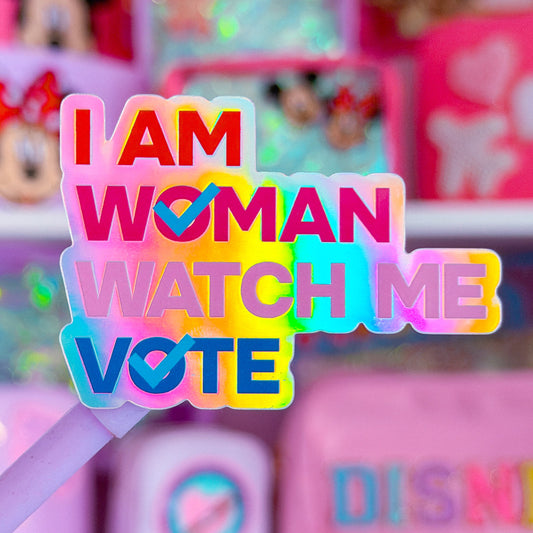 A close-up of a colorful text sign reading 'I AM WOMAN WATCH ME VOTE' held in front of shelves of pink and pastel stationery items, including Disney-themed products.
