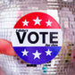 A round political sticker held by a finger, featuring a red, white, and blue design with the text 'I didn't VOTE for this' and white stars. The background shows a large, out-of-focus silver disco ball against a pink wall.