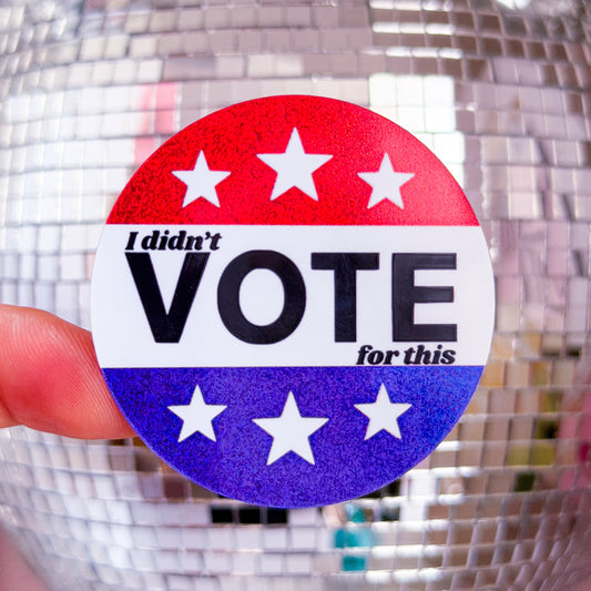A round political sticker held by a finger, featuring a red, white, and blue design with the text 'I didn't VOTE for this' and white stars. The background shows a large, out-of-focus silver disco ball against a pink wall.