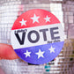 A round sticker held by a finger in front of a blurred disco ball. The sticker has a red, white, and blue design with white stars and the text 'I didn't VOTE for this'.