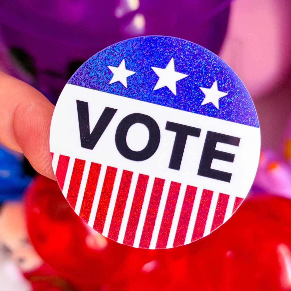 A hand holding a round 'VOTE' button badge with a blue glitter top half with white stars, a white middle band with black text, and a red and white striped bottom half, against a colorful blurred background.