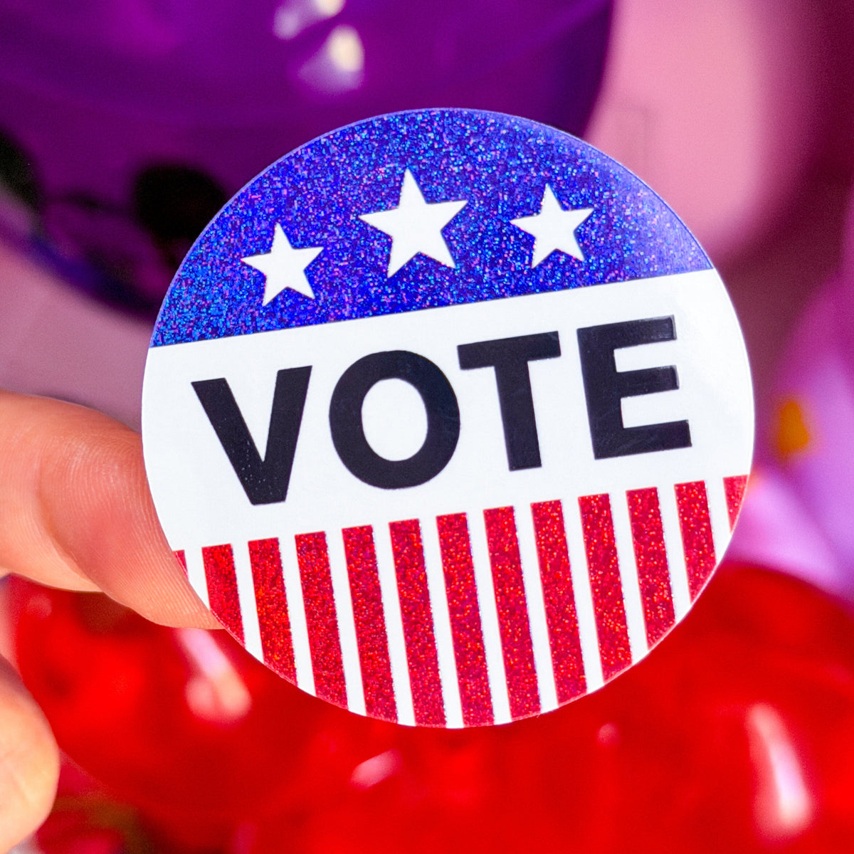 A hand holding a round glittery 'VOTE' button badge with a patriotic red, white, and blue design, set against a blurred background of purple and red balloons.